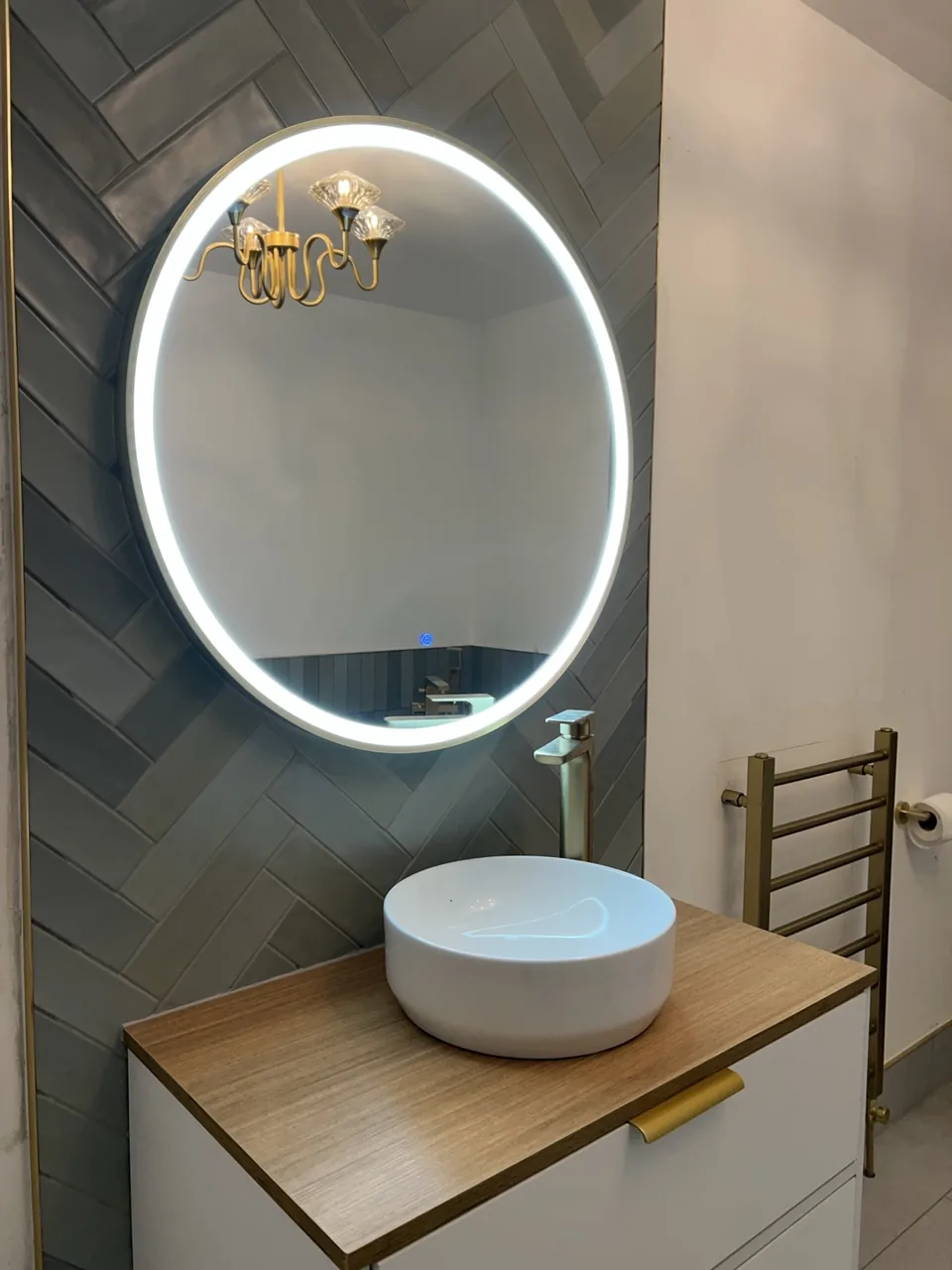 Herringbone tiled basin wall in a Limerick bathroom with an oval illuminated mirror, countertop basin and brass fittings